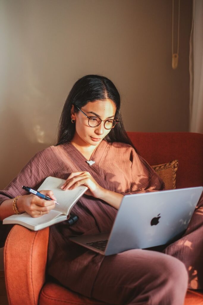 A woman with glasses sits in an armchair, using a laptop and taking notes in a notebook.