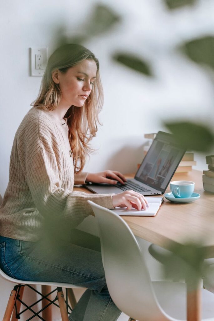 Side view of focused female freelancer reading notes in notepad while browsing netbook at table in room with blurred green leaves
